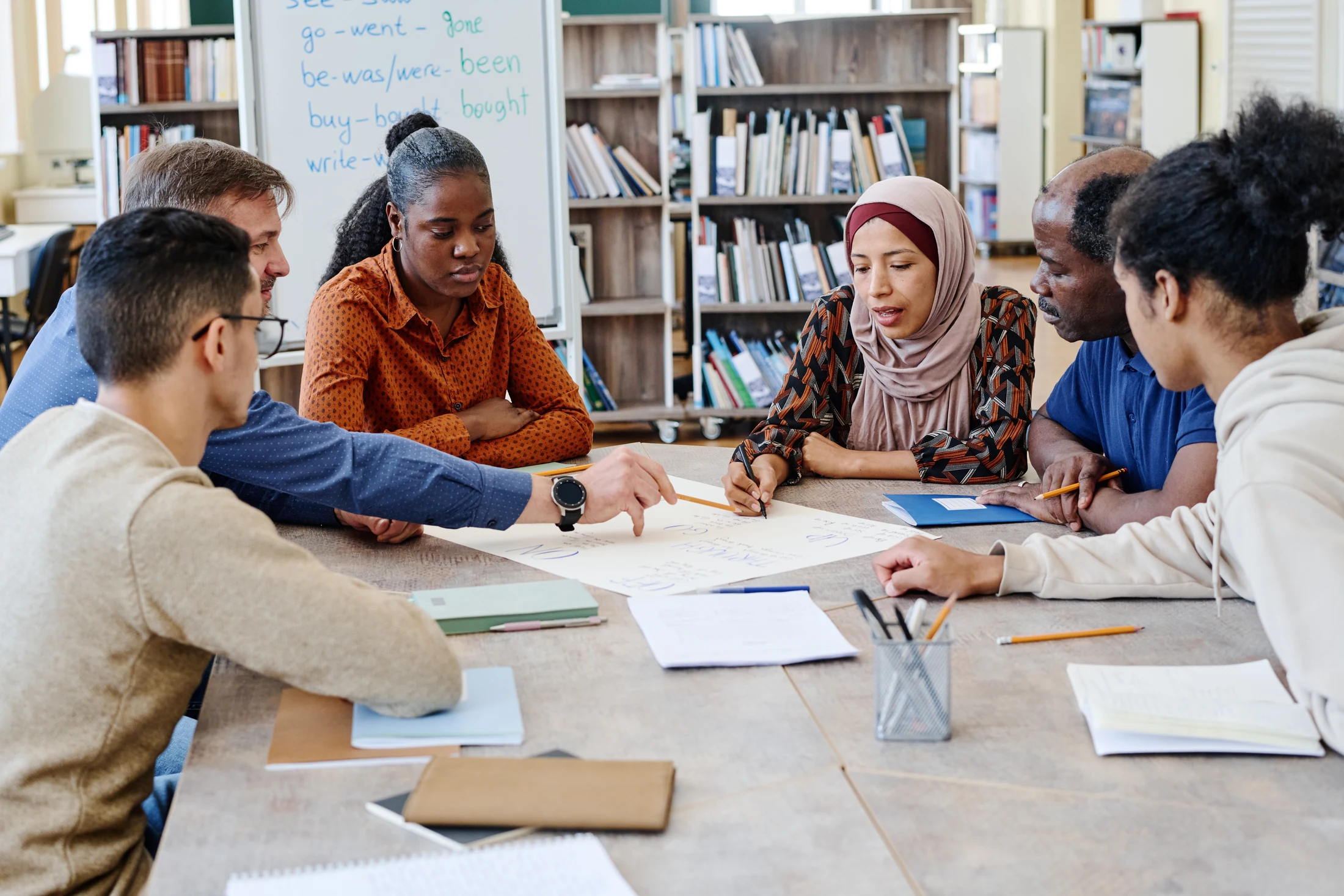 An instructor working through English verb forms with adult learners around a library table