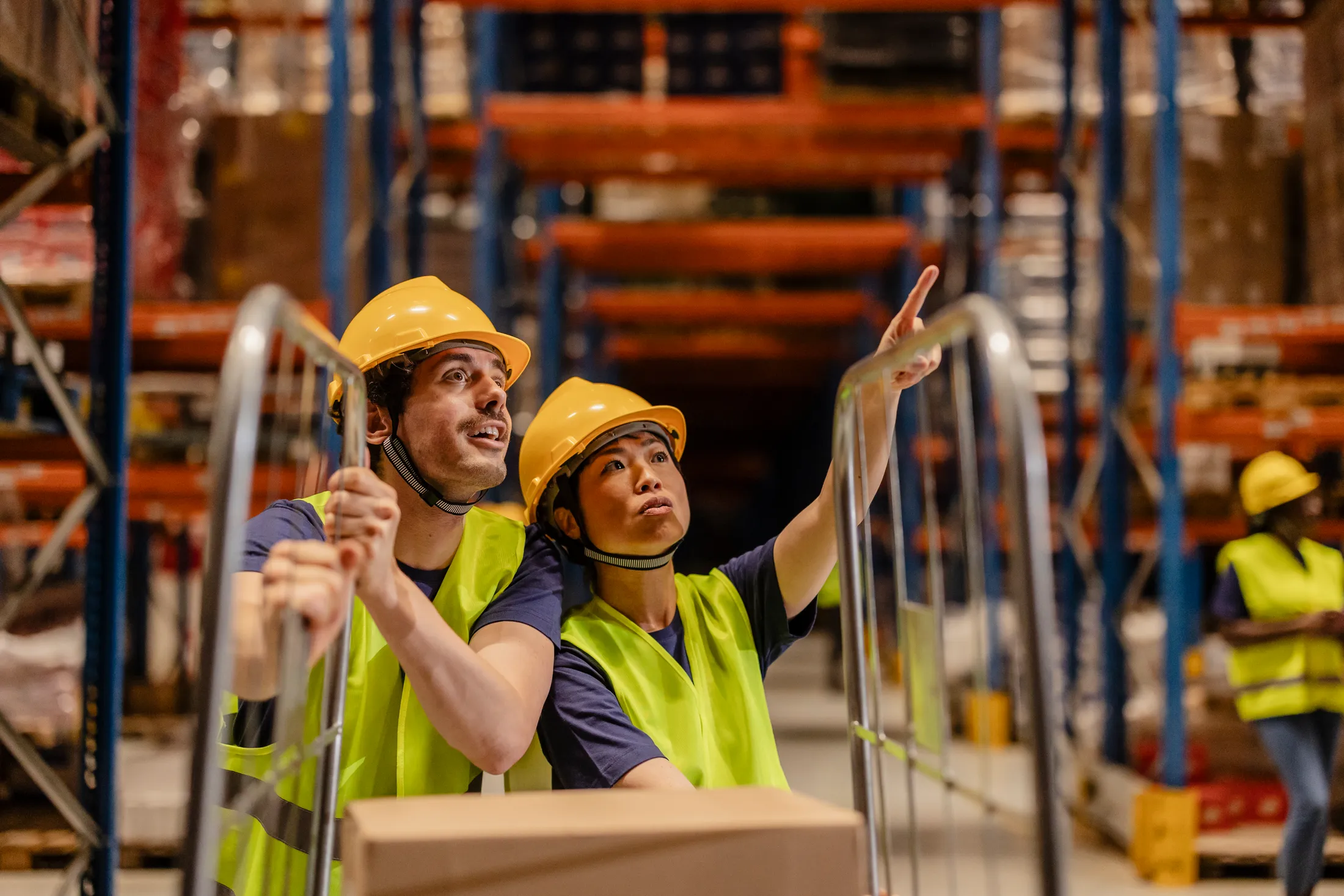 Two warehouse workers in hard hats and safety vests checking stock on high racking